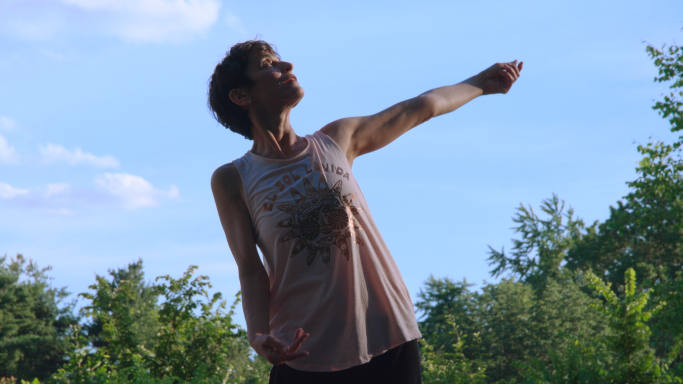 A wiry woman sporting short dark hair, sways to her right with her left arm lifted horizontally. She wears a light pink, no sleeved shirt that hits her thighs. She is wearing black pants. Her look is serene. She dances in front of a light blue sky. . 